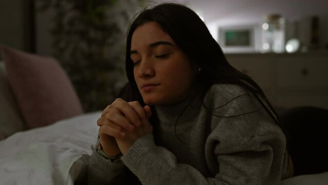 Hispanic teenager girl praying in a dimly lit bedroom at home, conveying a serene atmosphere.