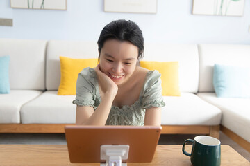An Asian woman is in the living room at home, holding a tablet and watching TV for entertainment and learning