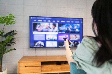 An Asian woman is watching TV in the living room at home, holding a TV remote control