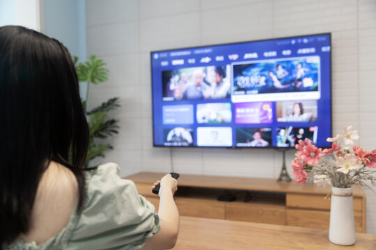 An Asian woman is watching TV in the living room at home, holding a TV remote control
