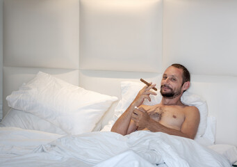Obraz na płótnie Canvas Caucasian bearded man sitting up in bed witch white sheets and pillows. Mid adult male is holding a cigar in one hand and a cigarette lighter in the other. The headboard of the bed is plain and white