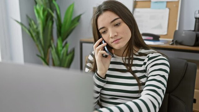 A young hispanic woman in an office converses on a phone, exuding professionalism and focus amidst technological and administrative surroundings.