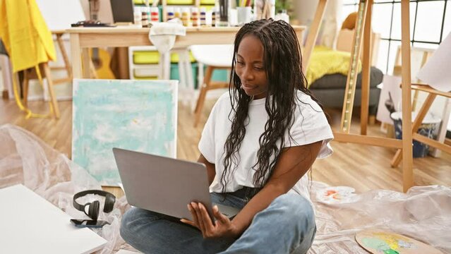 A young african american woman with curly hair working on a laptop in a creative art studio interior.