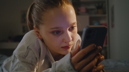 Close up of Caucasian teen lying on cozy bed in her comfort bedroom in the evening. Young girl uses her mobile phone to talk on video chat with friend. Attractive teenager spends weekend at home.