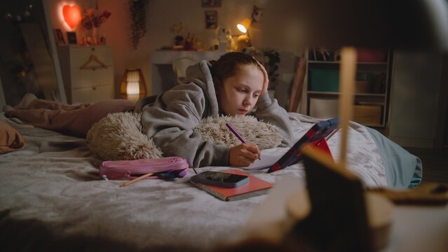 Teenage girl lies on bed at home, writes in notebook and does online homework for school using digital tablet computer. Young Caucasian girl spends time in cozy bedroom in daytime. Lifestyle concept.