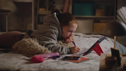 Fototapeta na wymiar Young girl lies on bed at home, writes in notebook and does online homework for school using tablet computer. Caucasian teenager spends time in cozy and comfort bedroom in daytime. Lifestyle concept.