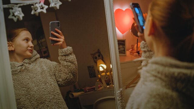 Young beautiful girl does makeup standing in front of the mirror in cozy bedroom, then takes photo using phone. Caucasian teenager spending leisure time at home in the evening. Concept of lifestyle.