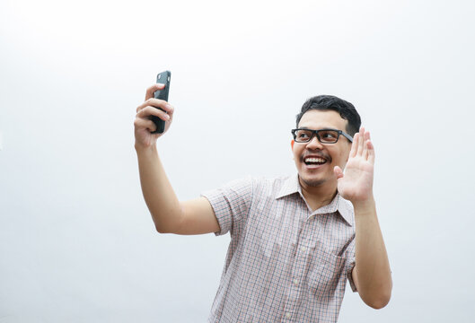 Handsome happy young Asian man wearing a white checkered shirt doing selfie shot on a mobile cell phone posts a photo on social network isolated over white background. People Lifestyle Concept