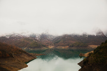 Lake surrounded by mountains with snowy tops and covered in fog