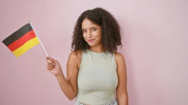 Radiant young hispanic woman, brimming with confidence, joyfully holding the german flag over an isolated pink background, expressing her patriotism with a captivating smile.