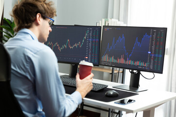 Working young business trader focusing on market stock graph data in two screens, holding coffee cup with back side view at modern office. Concept of analyzing dynamic financial exchange rate. Gusher. - obrazy, fototapety, plakaty