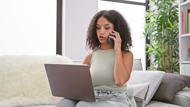 Hardworking hispanic woman in her prime, beautiful and serious, deftly juggling phone talk and laptop work, sitting focused on sofa. tech-savvy, yet homely indoor scene in living room.