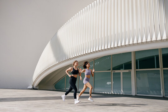Two active women athlete running side by side along an outdoor track on modern buildings background