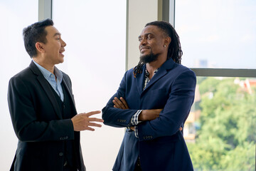 Asian businessman talking with African american businessman in seminar room near window and cityscape view.