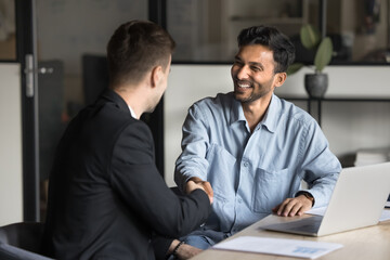 Cheerful multiethnic male business partners shaking hands at workplace table, smiling, laughing,...