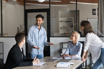 Happy young Indian manager speaking to colleagues, offering creative ideas for business project. Diverse team of coworkers brainstorming at job meeting, enjoying communication