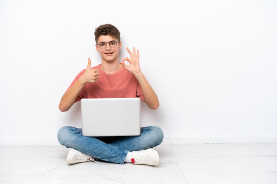 Teenager Russian man holding pc sitting on the floor isolated on white background showing ok sign and thumb up gesture
