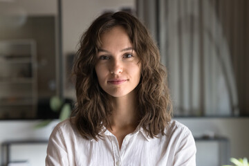 Positive beautiful business professional woman with wavy hair looking at camera standing in office, posing for portrait. Young entrepreneur, manager, employee head shot. Video call screen view - obrazy, fototapety, plakaty