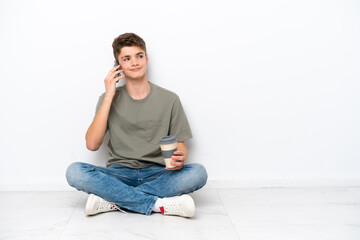 Teenager Russian man sitting on the floor isolated on white background holding coffee to take away and a mobile