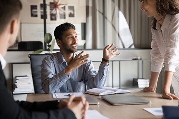 Positive inspired young Indian boss man talking to work colleagues at meeting table, brainstorming on marketing strategy, creative ideas, planning tasks, smiling. Business leader speaking to employees - obrazy, fototapety, plakaty