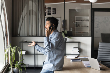 Serious young Indian business professional man in casual speaking on cellphone in office, leaning at workplace table looking at window away, talking to customer, moving hands. Side view