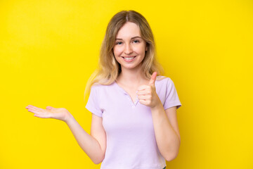 Blonde English young girl isolated on yellow background holding copyspace imaginary on the palm to insert an ad and with thumbs up