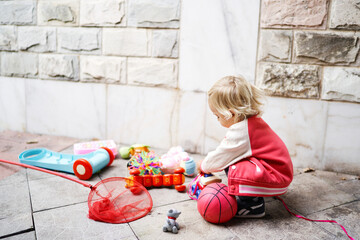 Little girl is playing with different colorful toys while squatting against a stone wall