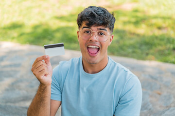 Young hispanic man holding a credit card at outdoors with surprise and shocked facial expression