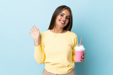 Young Uruguayan woman with strawberry milkshake isolated on blue background saluting with hand with...