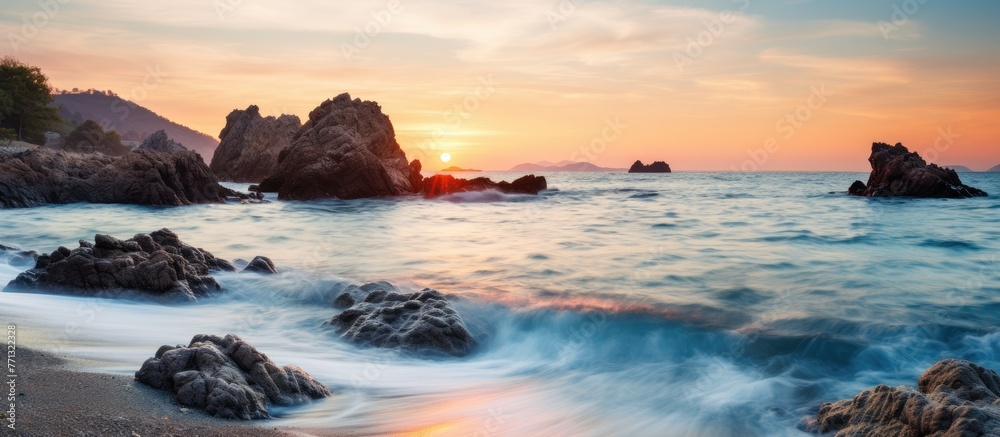 Aufkleber Sunset scenery over the ocean with rocks in the foreground and waves crashing on the shore, creating a serene natural setting