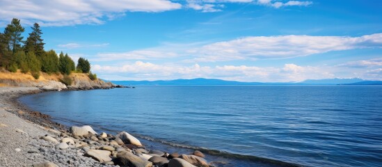 Rocky Puget Sound beach landscape with small rocks, calm sea, gentle waves, distant island, and clear blue sky with fluffy clouds