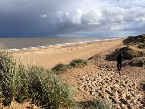 Man looking to ocean horizon beautiful sandy beach landscape grey stormy sky clouds and from grassy banks of sand dunes rolling waves on flat sea on Spring day in Winterton Norfolk East Anglia uk 