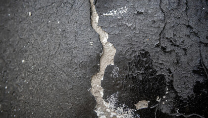 A close-up black and white shot of a cracked wall showing signs of deterioration and wear. The cracks cut through the concrete surface, creating a striking contrast between the dark shadows and light