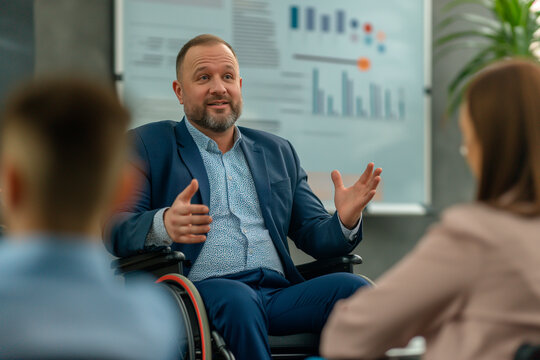 Disabled male business manager in wheelchair giving presentation for other people at meeting on chartboard