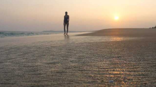 Young woman in swimsuit walking on sand beach near ocean at sunset. Beautiful young girl going on sea shore with waves and enjoying vacation. Concept of relaxing on summer holiday. Slow motion
