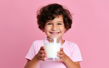 A smiling young child holds a glass of milk against a pink background