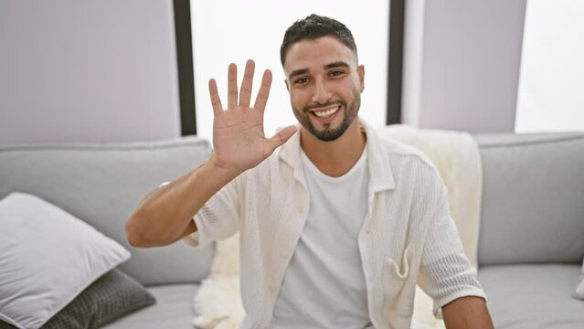 Cheerful young arab man sitting on sofa at home, ecstatically counting five on fingers, pointing up with a happy smile