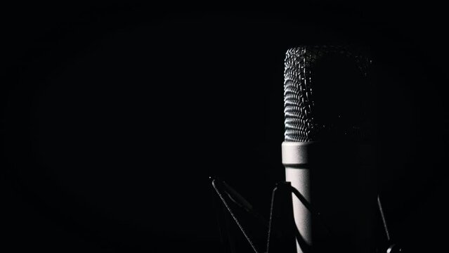 A studio microphone rotates on a black background in backlight close-up. Condenser microphone with chrome grid on surface. Concept recording studio, voice, podcast, karaoke, audiobook. Copy space