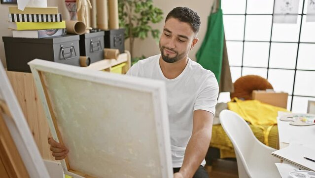 Smiling bearded man holds canvas in creative studio showcasing artistic casual interior