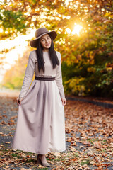 A fashionable woman with wide-brimmed hat smiling amidst autumn's glow in the park and posing for camera