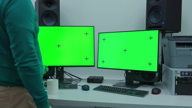 A man step in the scene with two computer monitors are on a desk with a green screen and add a milk to a coffee cup stands on a table. The desk is cluttered with various items such as a keyboard, mous