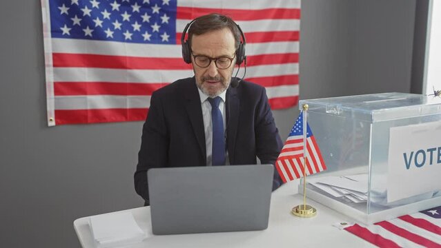Mature man with headphones using laptop in american voting center with us flag and ballot box.