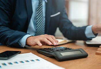 executive, strategy, payment, calculate, chart, document, graph, statistics, bookkeeper, management. A man in a suit is using a calculator on a desk. The desk is cluttered with papers and a phone.