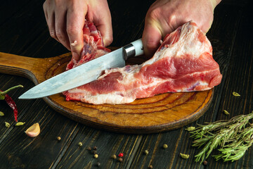 A butcher uses a knife to cut raw meat on a kitchen cutting board before preparing a meat dish for...