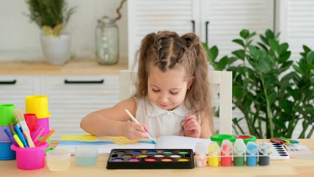 a happy and joyful child paints at a table in a bright room, a little girl learns to draw with a brush and gouache, a drawing lesson