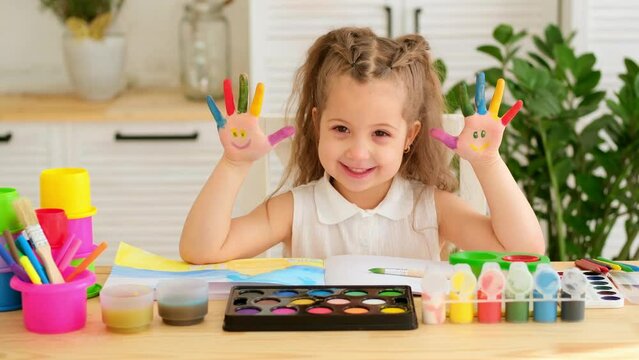 a happy and joyful child paints at the table or dabbles in painting and getting his hands dirty, a little girl learns to draw with a brush and gouache, a drawing lesson