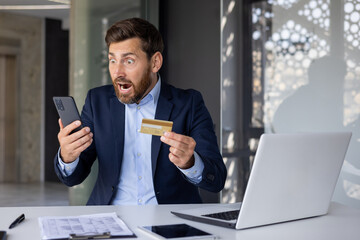 Shocked young man in business suit sitting at desk in office, holding credit card in hand and looking surprised at mobile phone screen - obrazy, fototapety, plakaty