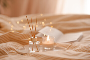 Fototapeta na wymiar Liquid home perfume in glass bottle with bamboo sticks and open paper book with scented candle on ceramic tray in bed over glowing lights. Cozy atmosphere.