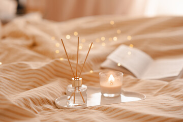 Liquid home perfume in glass bottle and scented candle with bamboo sticks and open paper book on ceramic tray in bed over glowing lights. Cozy atmosphere.