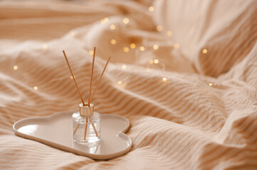 Liquid home perfume in glass bottle with bamboo sticks on ceramic tray in bed over glowing lights. Cozy atmosphere.